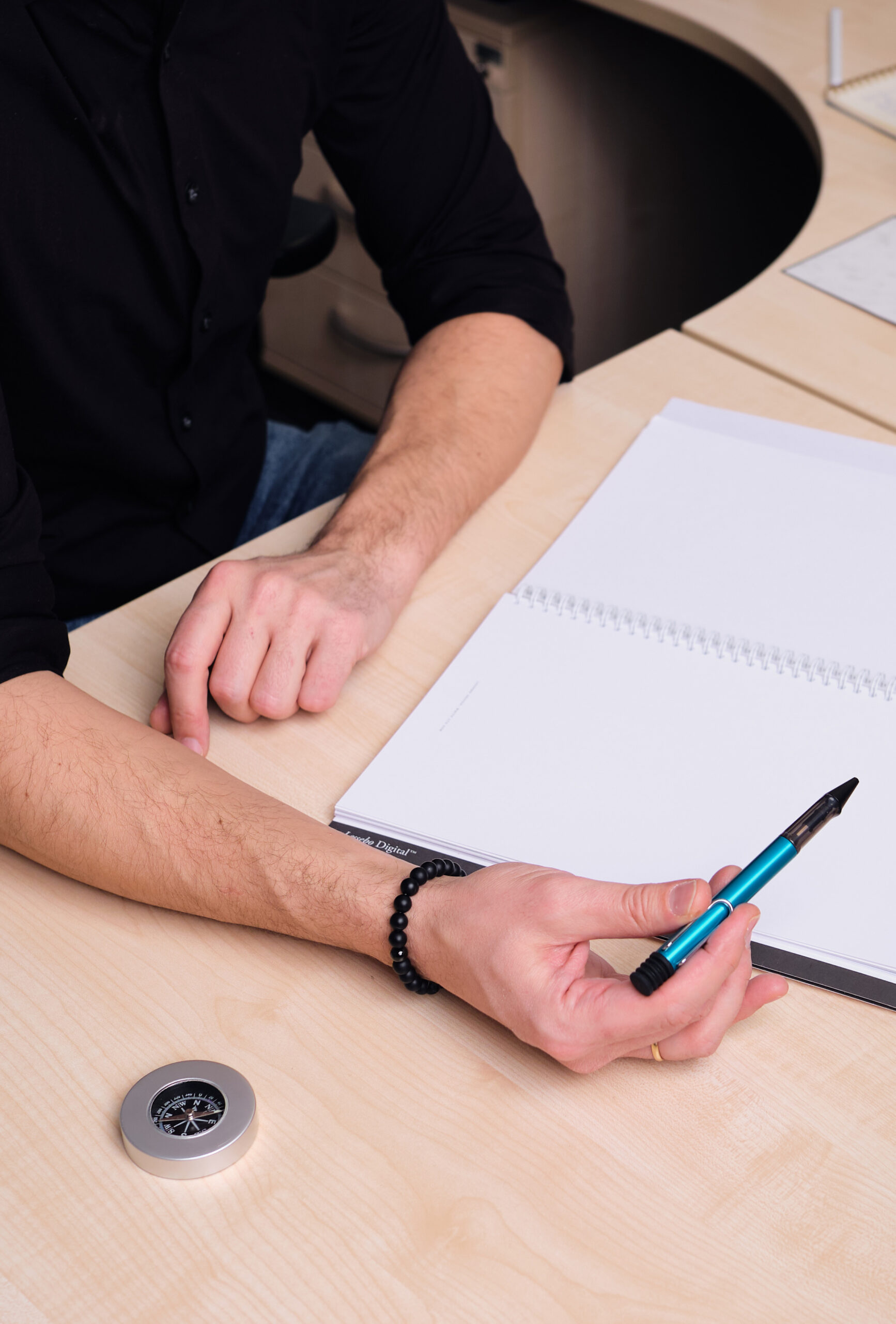 Eine Hand hält einen Stift über ein weißes Blatt Papier. Auf dem Tisch steht ein Kompass.